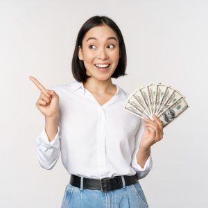 Smiling young modern asian woman, pointing at banner advertisement, holding cash money dollars, standing over white background.